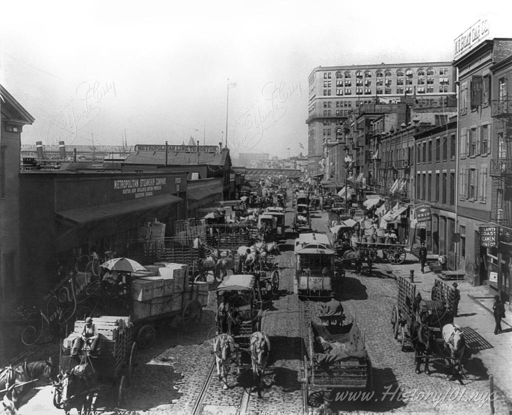 View along waterfront on West Street with many freight wagons, street cars and the buzz of daily activity.