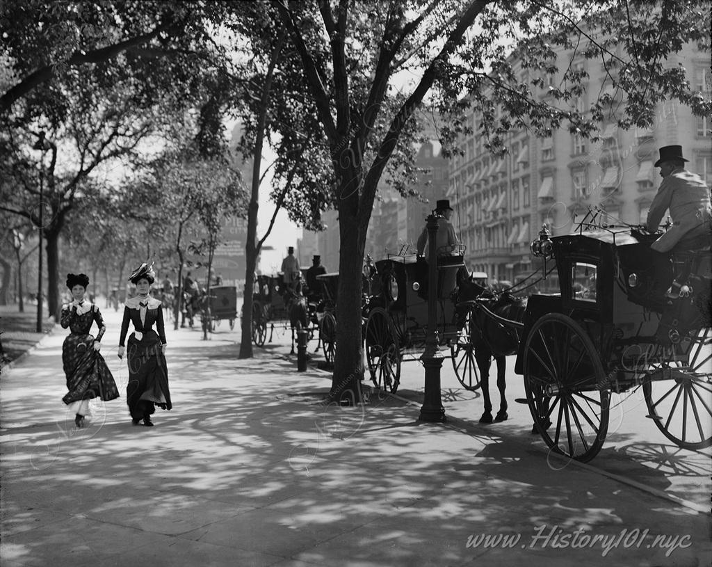 Two women passing a row of horse-drawn carriages as they walk alongside Madison Square Park.