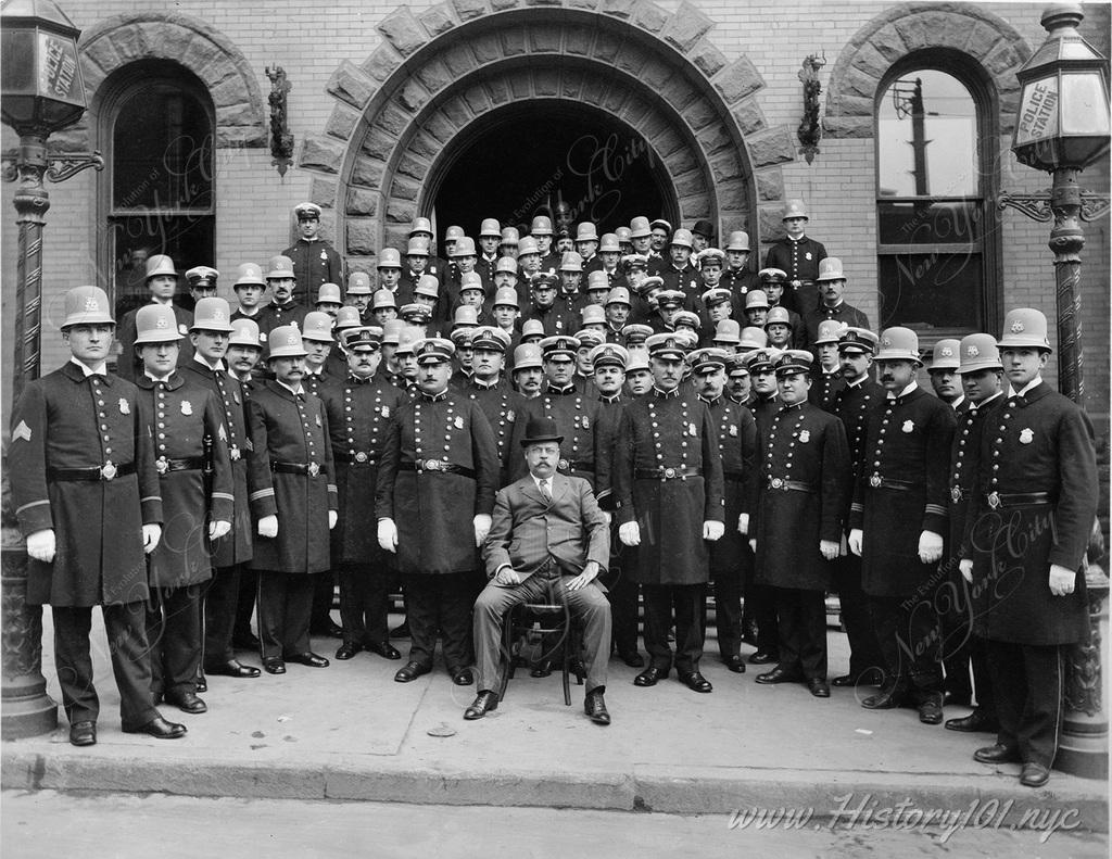 A group of New York City police officers pose together in front of a precinct.