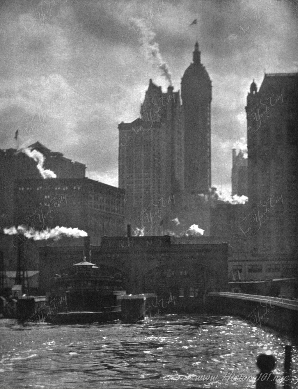 Dramatic image of Manhattan's downtown skyline behind the East River shows steam rising from various smoke stacks. Photographed by Alfred Stieglitz.