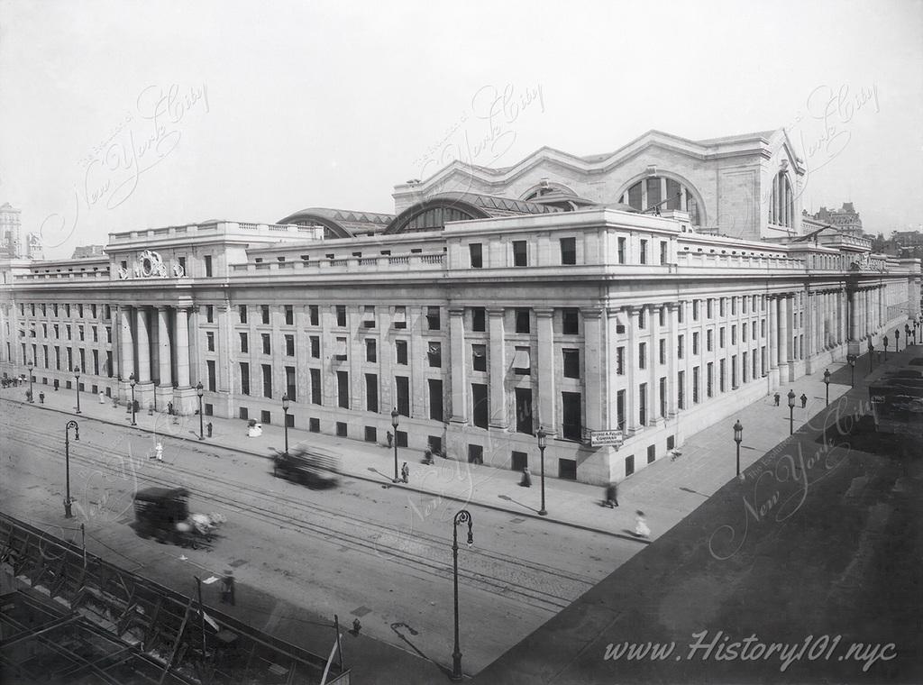Photograph from an elevated perspective showing 32nd and 33rd Street and 7th Avenue and the facade of Pennsylvania Station as horses and carriages pass by.