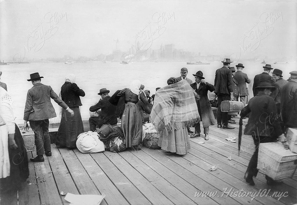 Photograph of immigrants waiting to be transferred at Ellis Island on October 30, 1912. Manhattan's downtown skyline is visible in the distance.