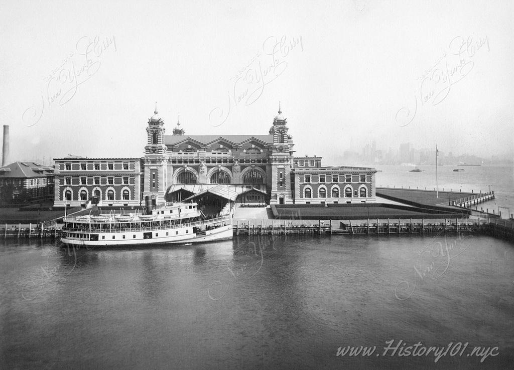 An excellent overhead view of the front facade of Ellis Island's Immigration Station with a boat docked in front.