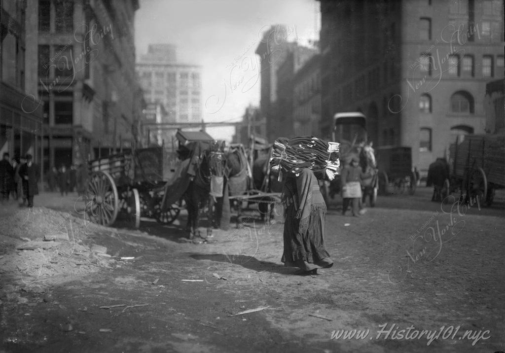 A senior citizen is doubled over as she carries a stack of textiles across Lafayette Street below Astor Place.