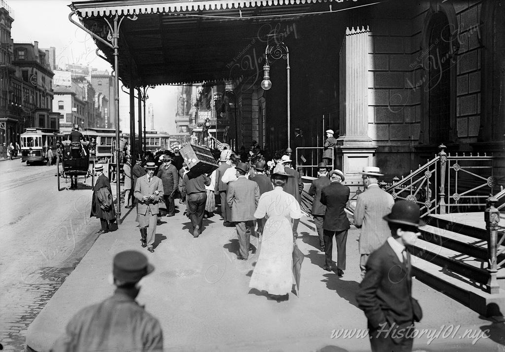 A crowd of pedestrians pass the entrance of Grand Central Terminal with horse carriages and trolleys visible on the street.