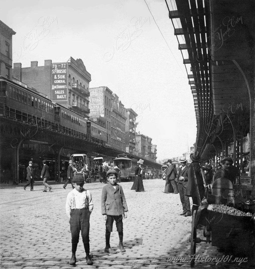 Photograph of a busy scene on the Bowery as pedestrians make their way across the street and under the elevated overpass.