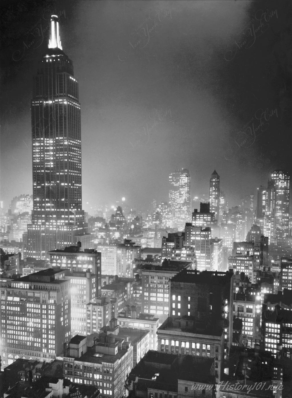 Photograph of the Empire State Building and surrounding skyscrapers illuminated at night.