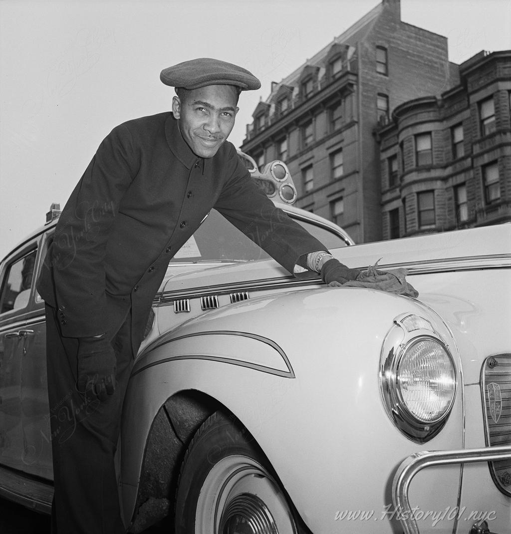 Photograph of a taxi driver polishing the hood of his car in front of a row of brownstones.