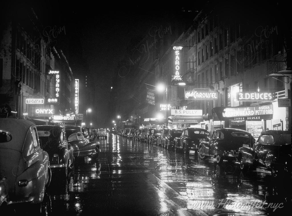 Rows of parked cars reflect the neon lights of the bars and clubs on a rain-soaked 52nd Street.