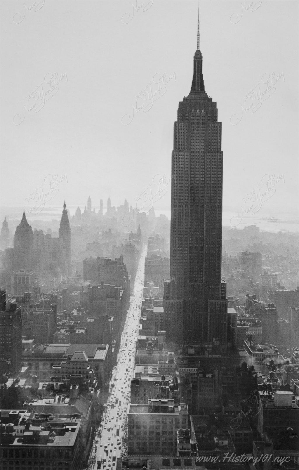 An elevated perspective of Midtown Manhattan's famous skyscrapers with the Empire State Building casting a long shadow as the sun illuminates 5th Avenue.