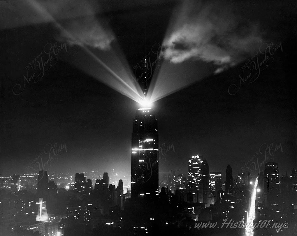Photograph shows beacon lights beaming brightly from atop the Empire State Building at night illuminating the clouds in the sky.