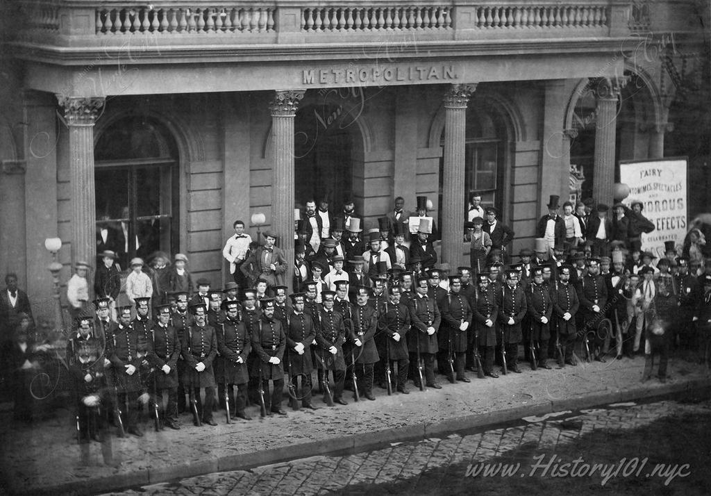 Group portrait of policemen, standing in rows in front of a Metropolitan building with their hands crossed in front of them and guns resting at their sides.