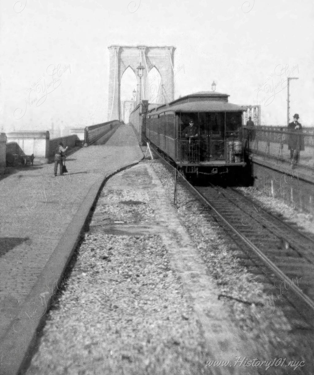 Photograph of a the trolley tracks on the Brooklyn Bridge. A city worker is seen cleaning the platform on the left while commuters are seen walking on the right.
