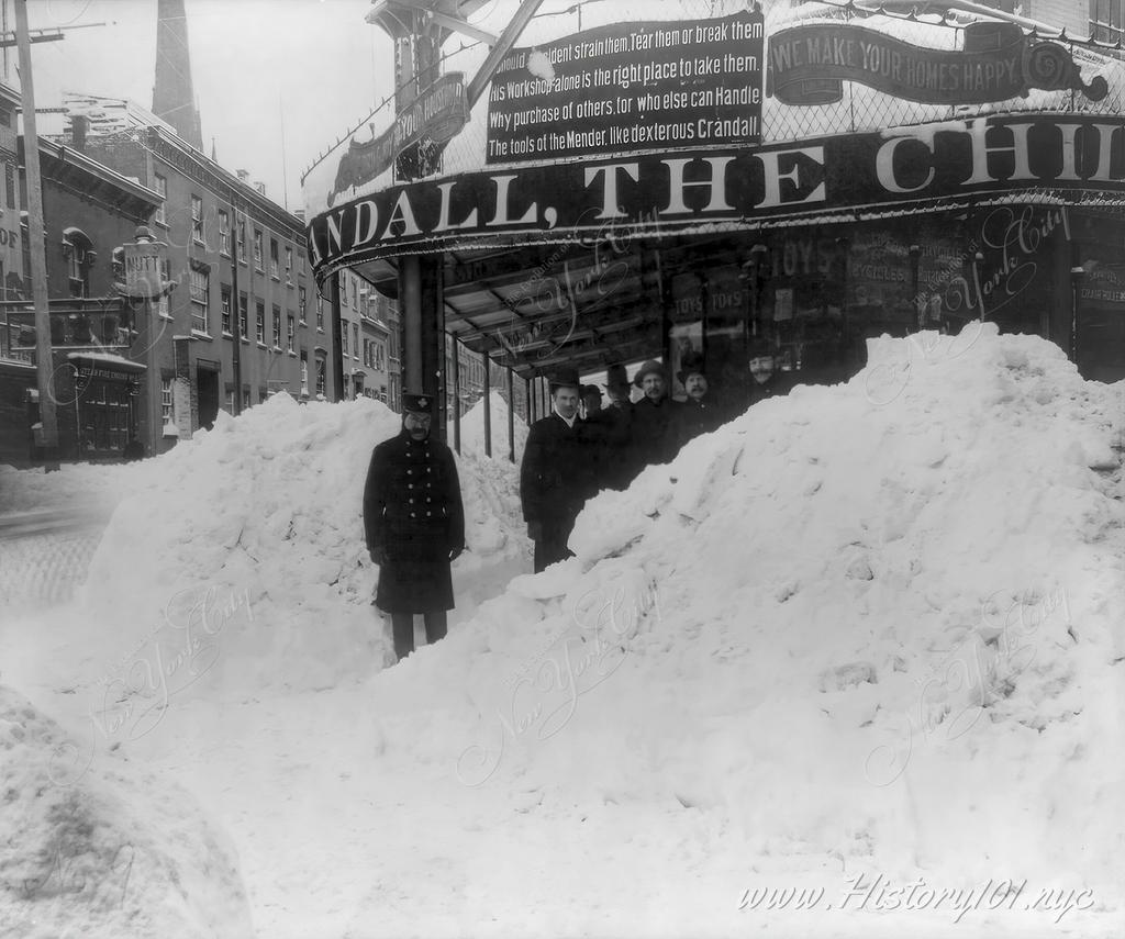 Photograph shows piles of snow taller than a group of men posing in front of a storefront during the Blizzard of 1888.