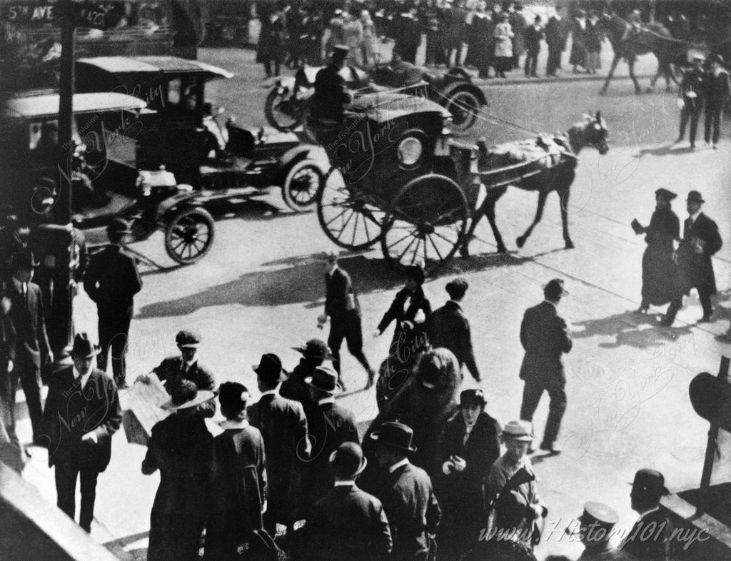 Photograph shows a horse-drawn carriage and automobiles on a city street in strong sunlight.