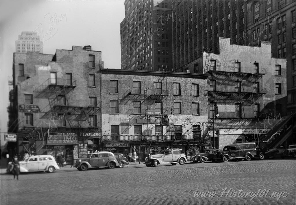 Photograph of cars parked in front of the buildings in facing Battery Park, between Washington And Greenwich Streets.