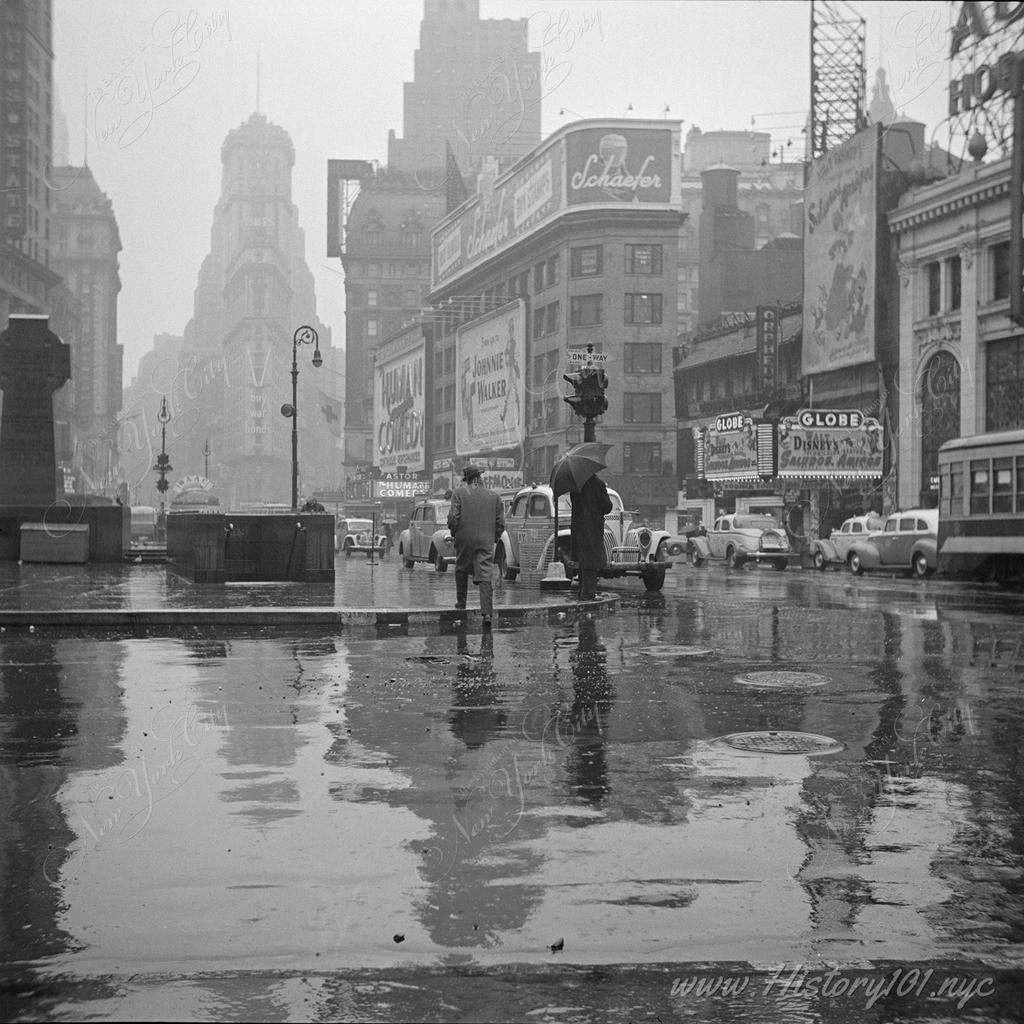 Photograph of wet street and pavements at Time Square with the Times Building barely visible through the rain.