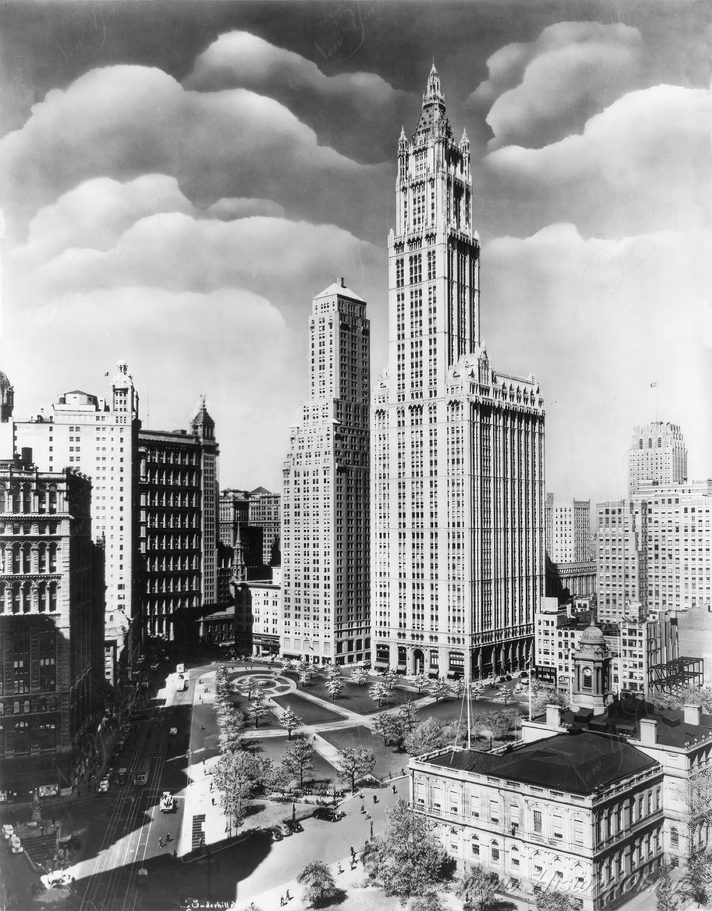 A bird's eye view of the Woolworth Building towering above City Hall Park on a partly cloudy day.