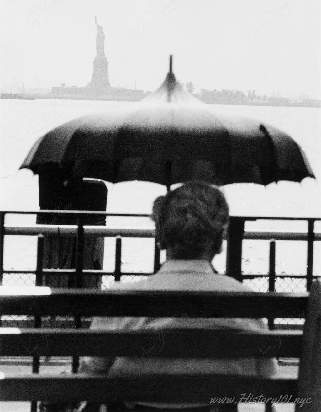 Photograph of a man holding an open umbrella, sitting on a dock with a view of the Statue of Liberty across the harbor.