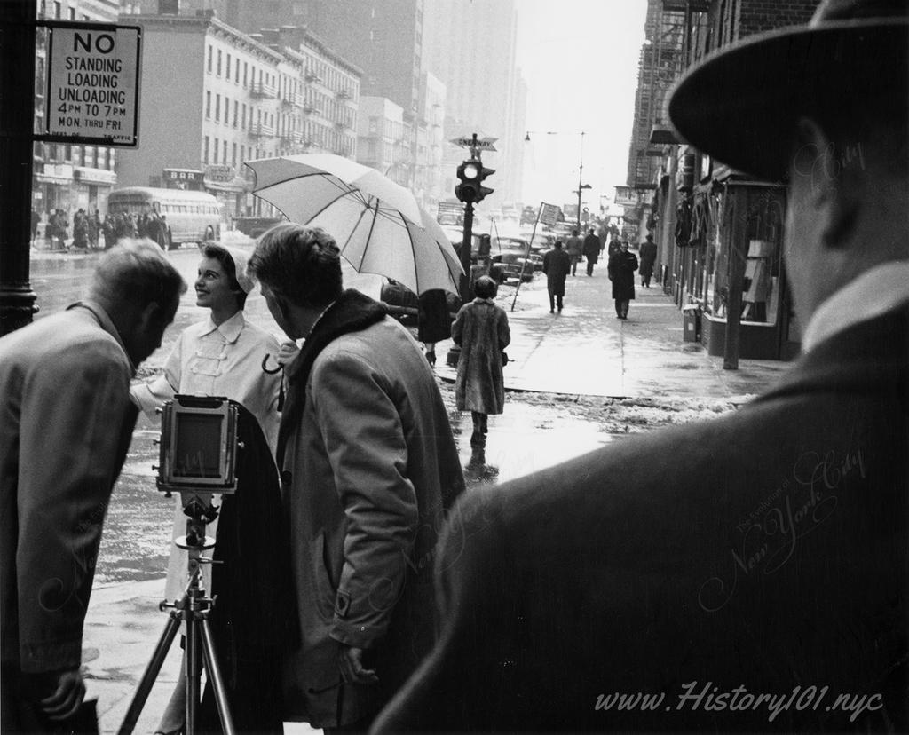 Photograph shows a woman being photographed on a street corner. Two men, one holding an umbrella stand behind the camera which is on a tripod. 