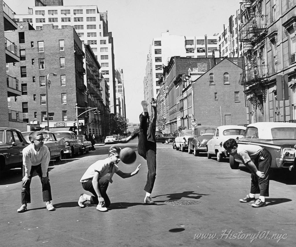 Photograph of a local game of kickball being played in the middle of MacDougal Street in Manhattan.
