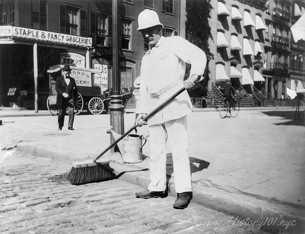 Photograph of a city worker sweeping the street in front of a corner grocery store.