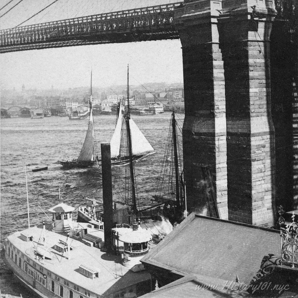Photograph of East River boats passing underneath the Brooklyn Bridge overpass.