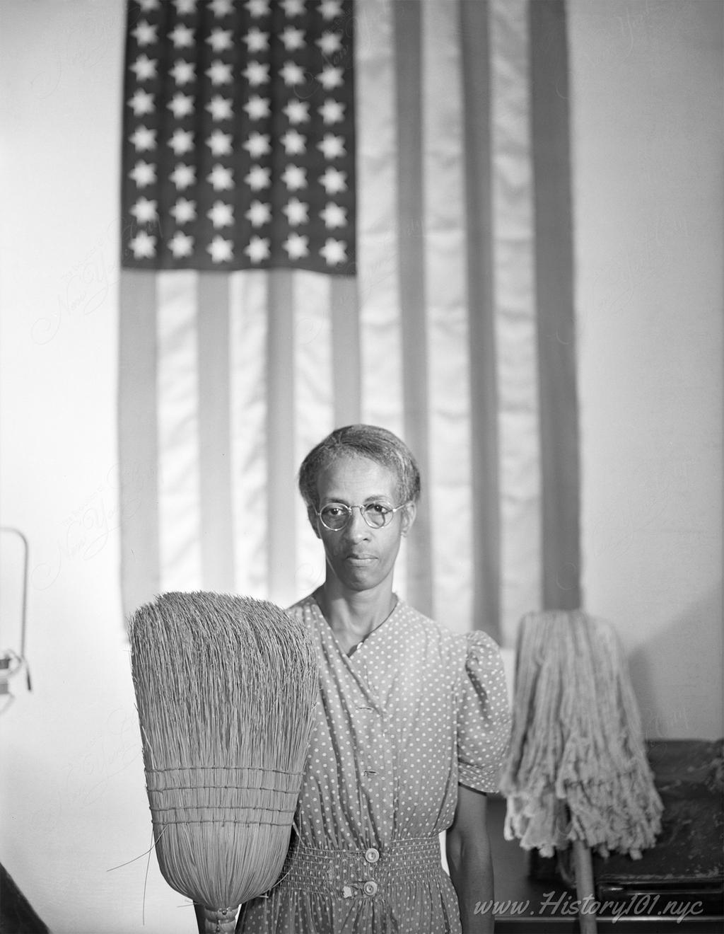 Portrait of Ella Watson standing in front of the American flag with mop and broom.