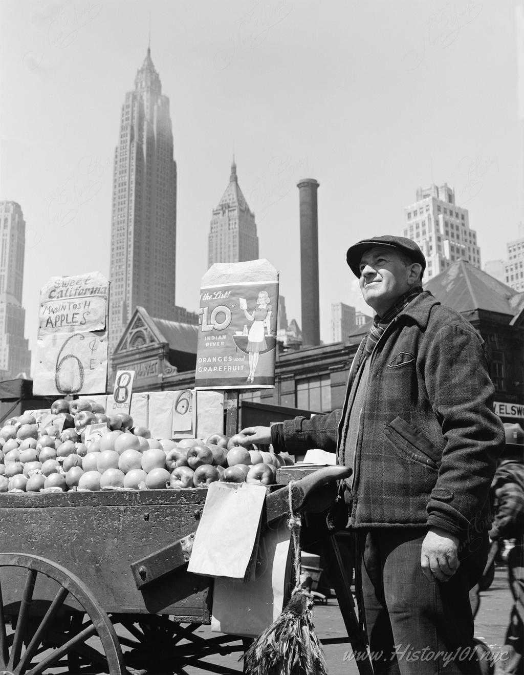 Photograph of a pushcart fruit vendor at Fulton Fish Market with downtown Manhattan skyscrapers visible in the background.