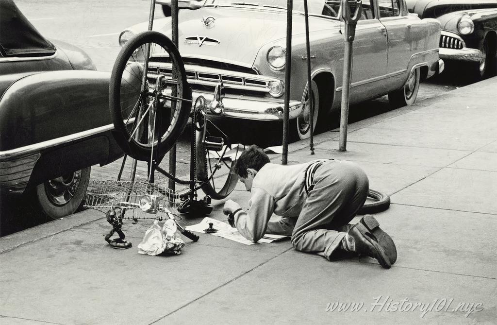 Photograph of a man crouched on a sidewalk working on a bicycle, which is turned upside down in front of him.