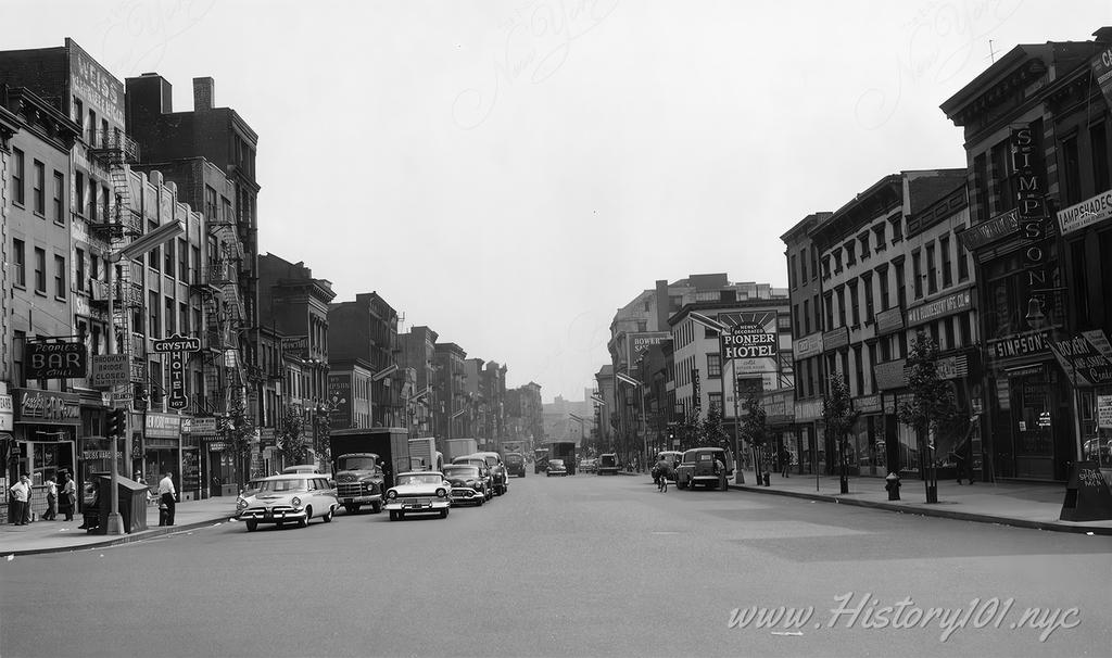 Photograph of The Bowery and Delancey Street showing cars at a stoplight and signs for the People's Bar, and the Crystal Hotel and Pioneer Hotel in distance.