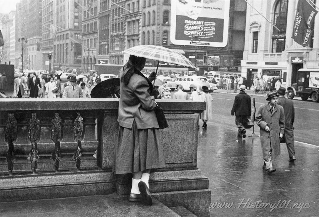 Photograph of a woman holding an umbrella and leaning on a bannister against a bustling backdrop of pedestrians, traffic, and storefronts.