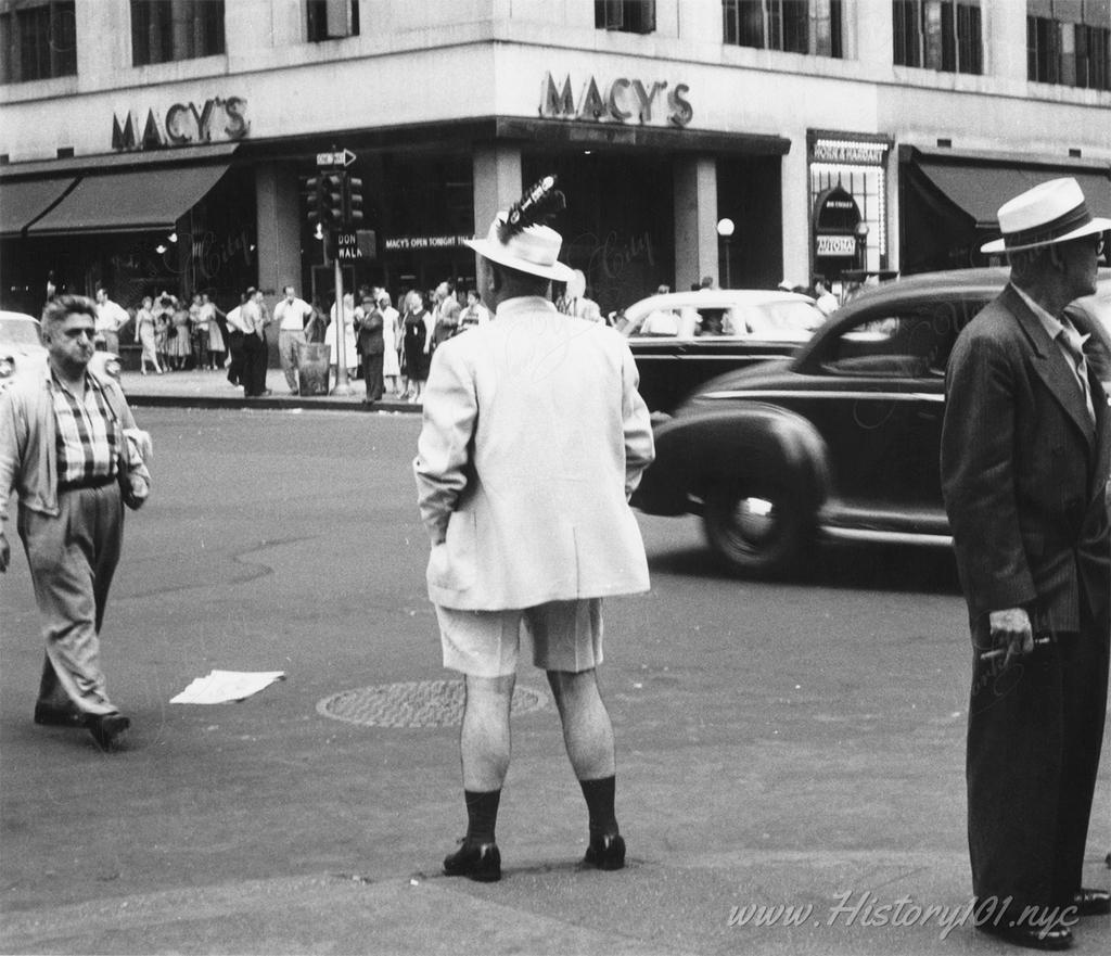 Photograph of a man in shorts with feathered cap, standing on a street curb with Macy's department store in the background.