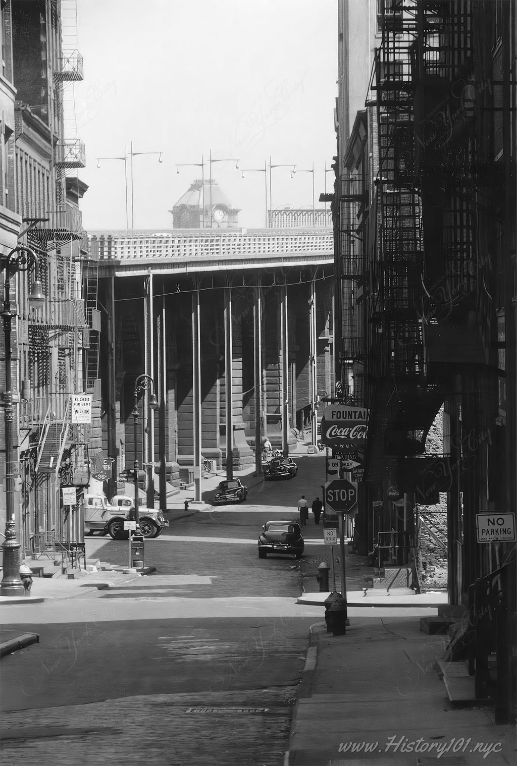 Photograph shows cars parked along empty street and street signs with the Brooklyn Bridge overpass in the background.