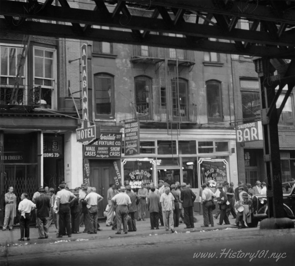 Photograph shows men gathered on sidewalk in front of the Alabama Hotel and Skid Row Bar & Grill in downtown Manhattan.
