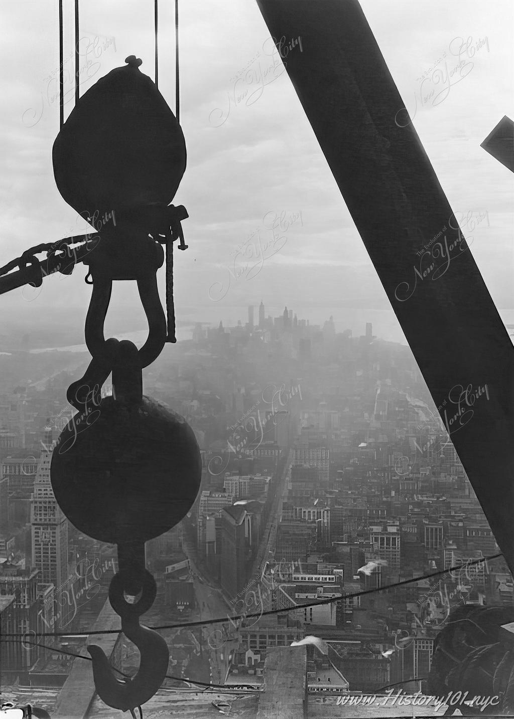 Aerial photograph taken from inside the growing structure of the Empire State Building. An epic view of downtown Manhattan is framed by the silhouette of a massive crane hook and steel beam.