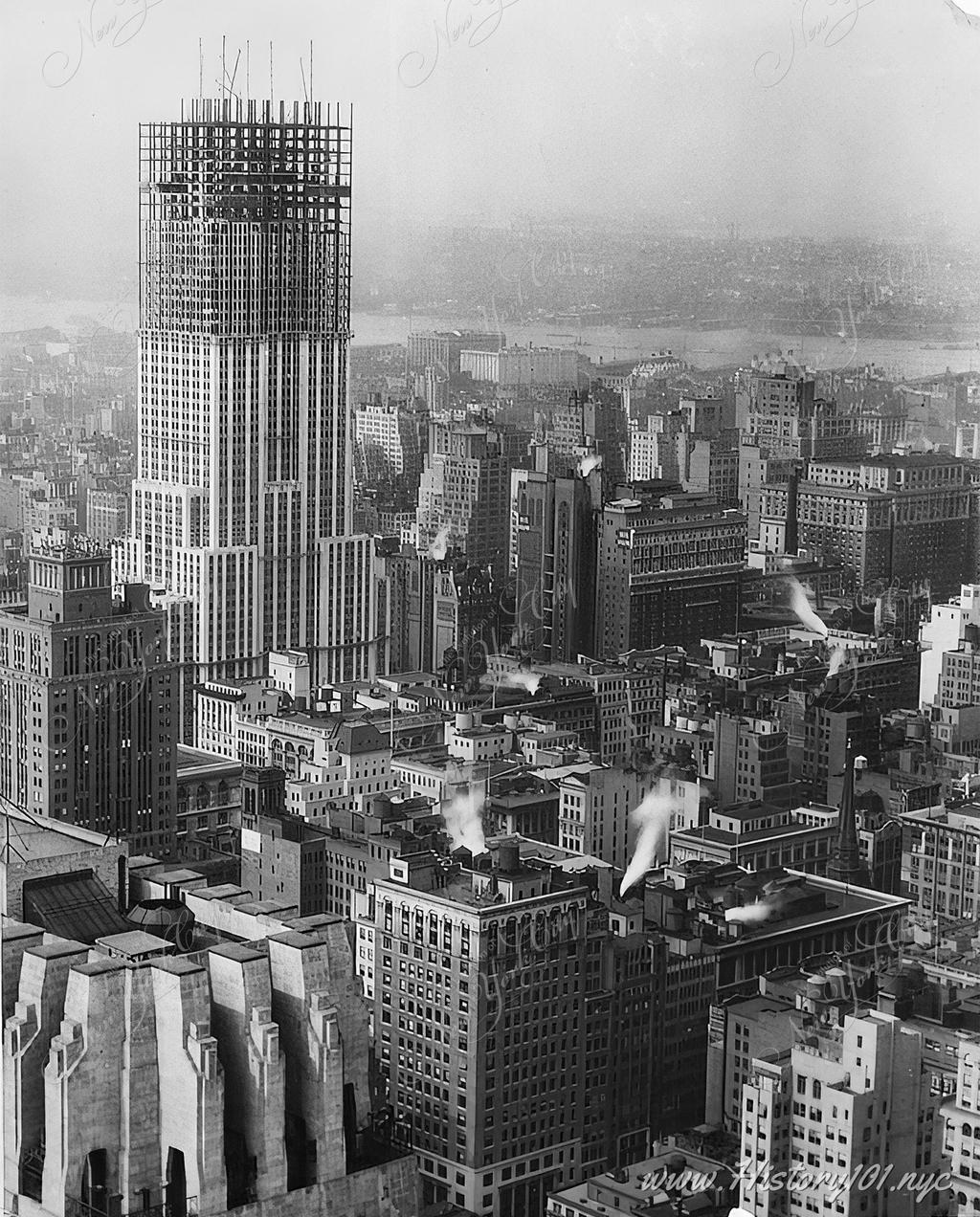 Bird's eye view of midtown Manhattan and the Empire State Building, about 75% complete with half of its steel structure visible.