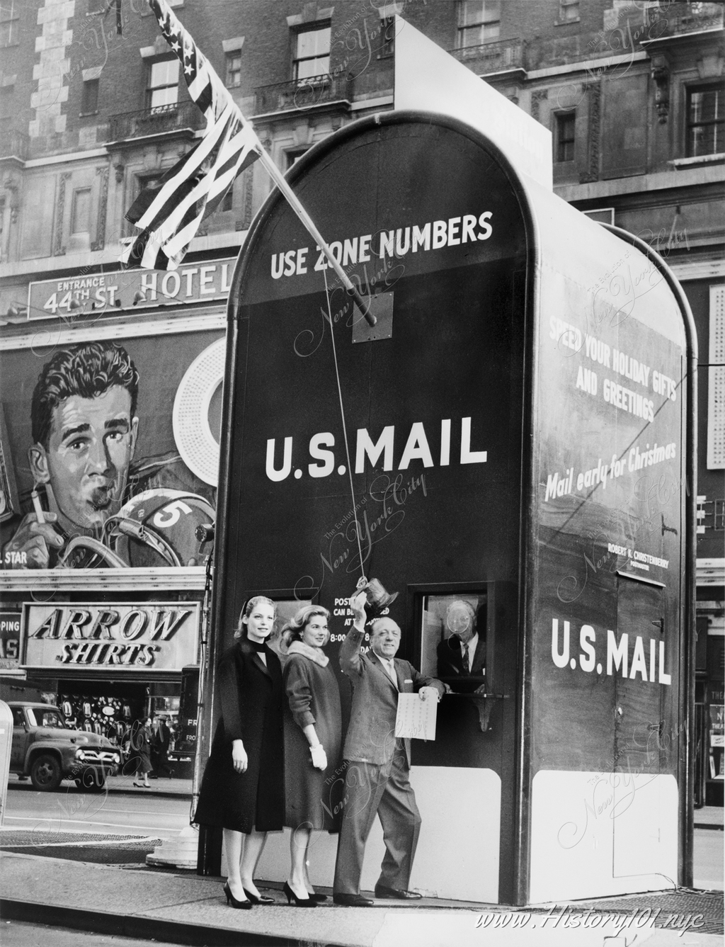 Actresses Millette Alexander and Louise King, and nightclub entertainer Ted Lewis, stand outside a giant mailbox stamp selling booth in Times Square, New York City, while Assistant Postmaster Aquiline F. Weierich dispenses stamps from inside booth